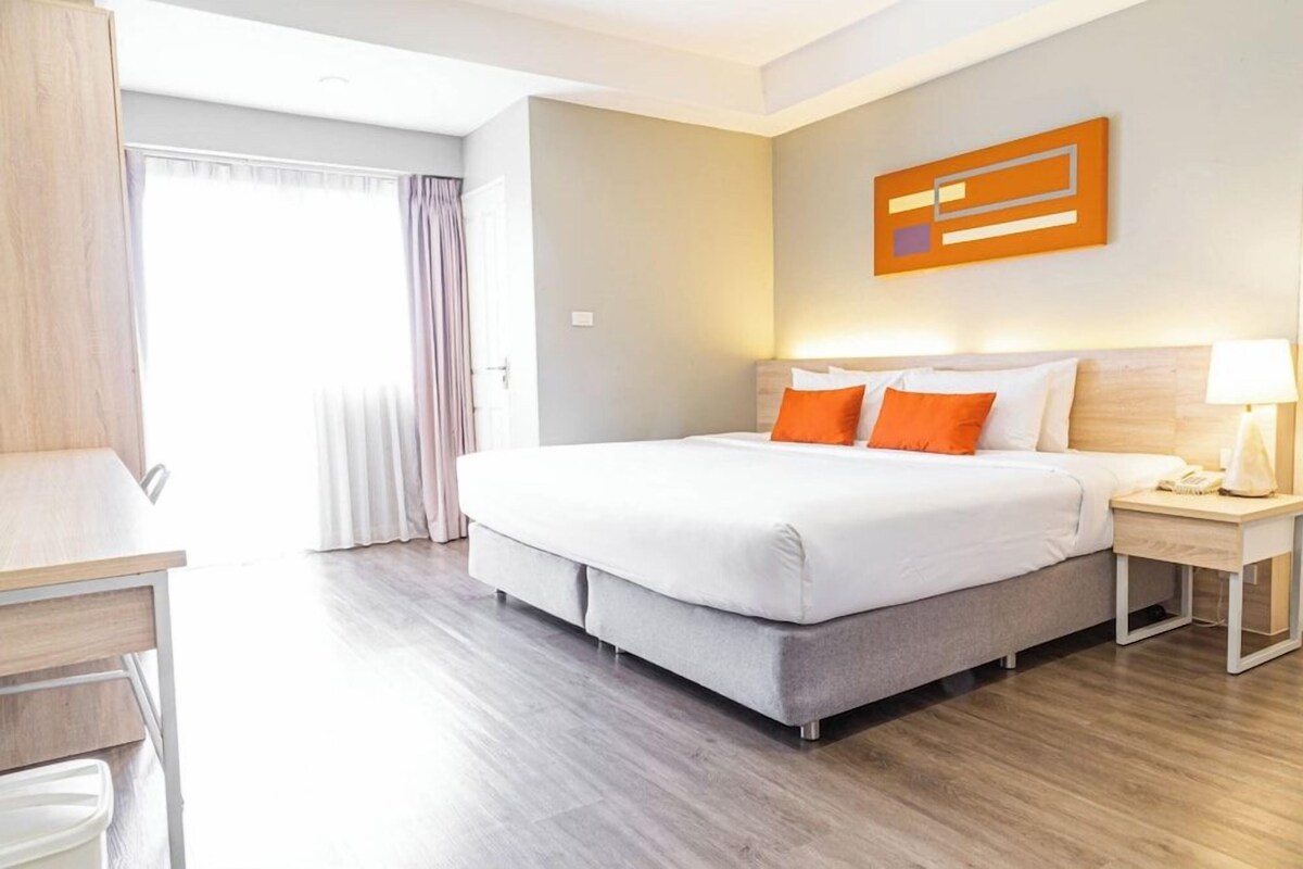 Habitación de hotel con cama, mesita de noche, lámpara y vistas al exterior.