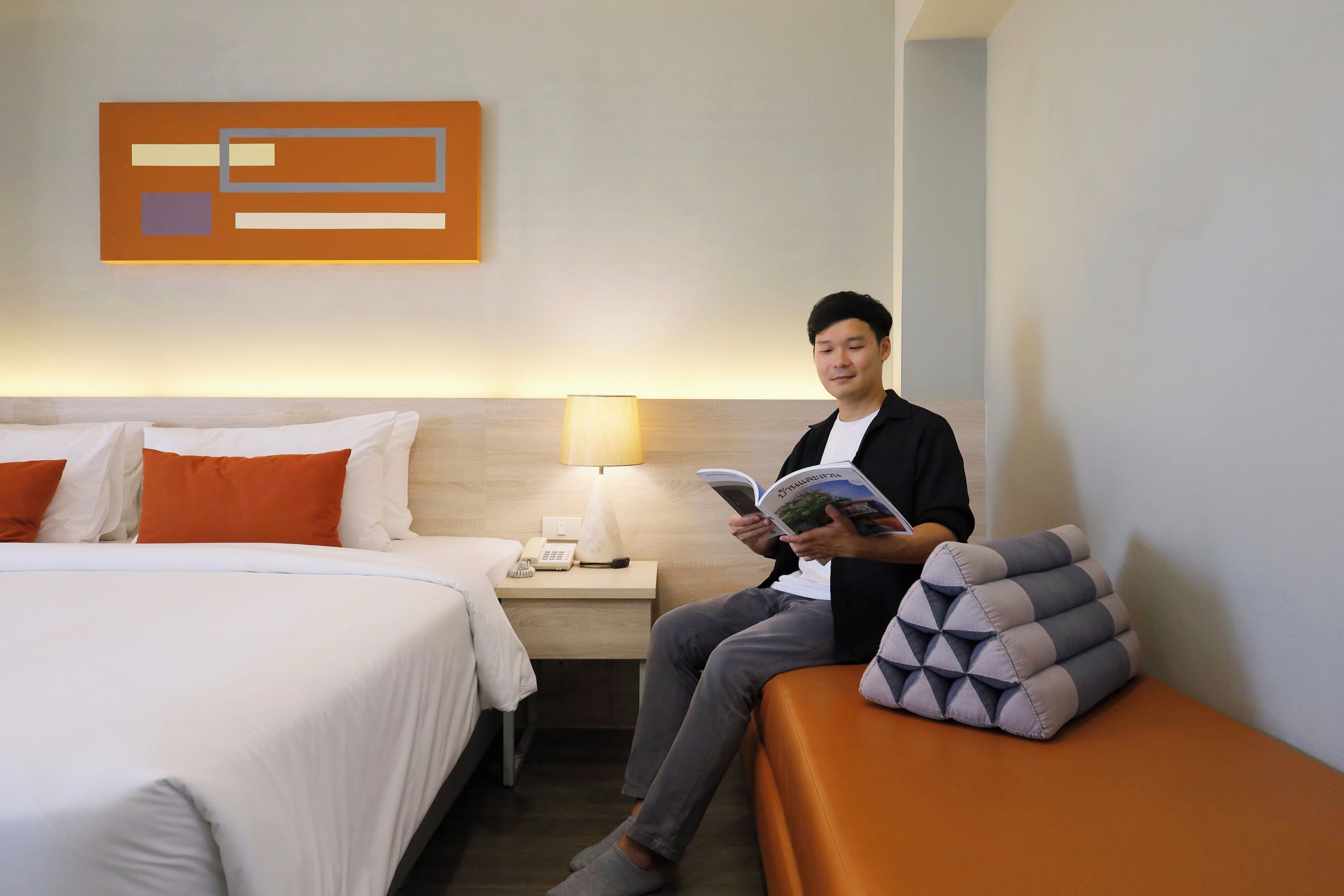 A hotel room with a bed, a bedside table with a lamp, and a person reading a book.