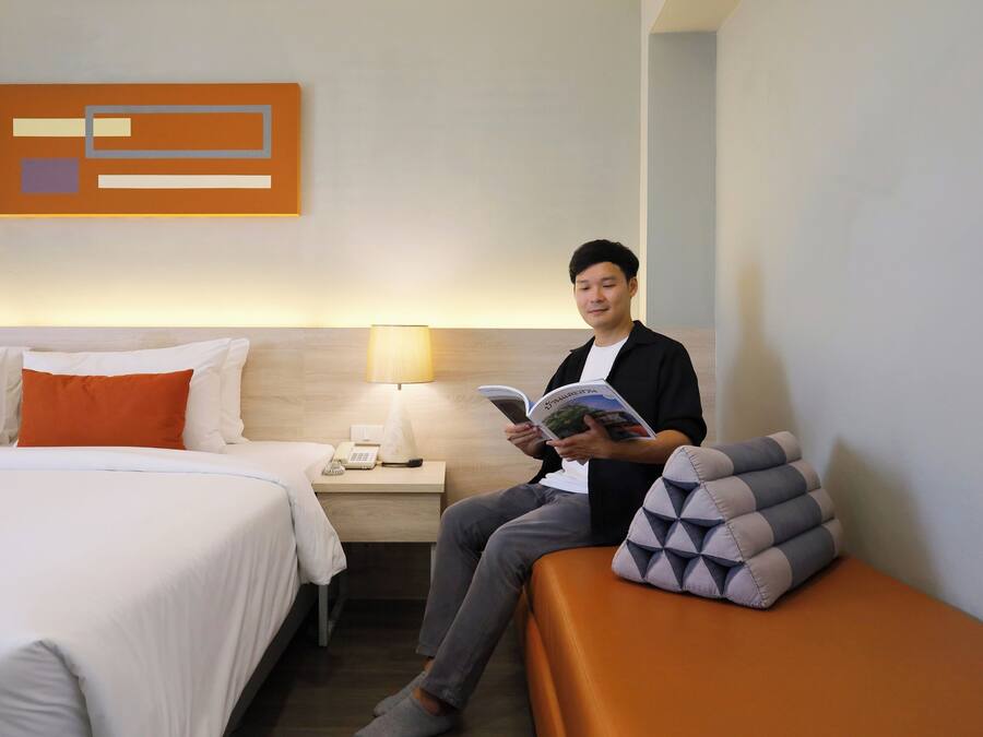 A hotel room with a bed, a bedside table with a lamp, and a person reading a book.