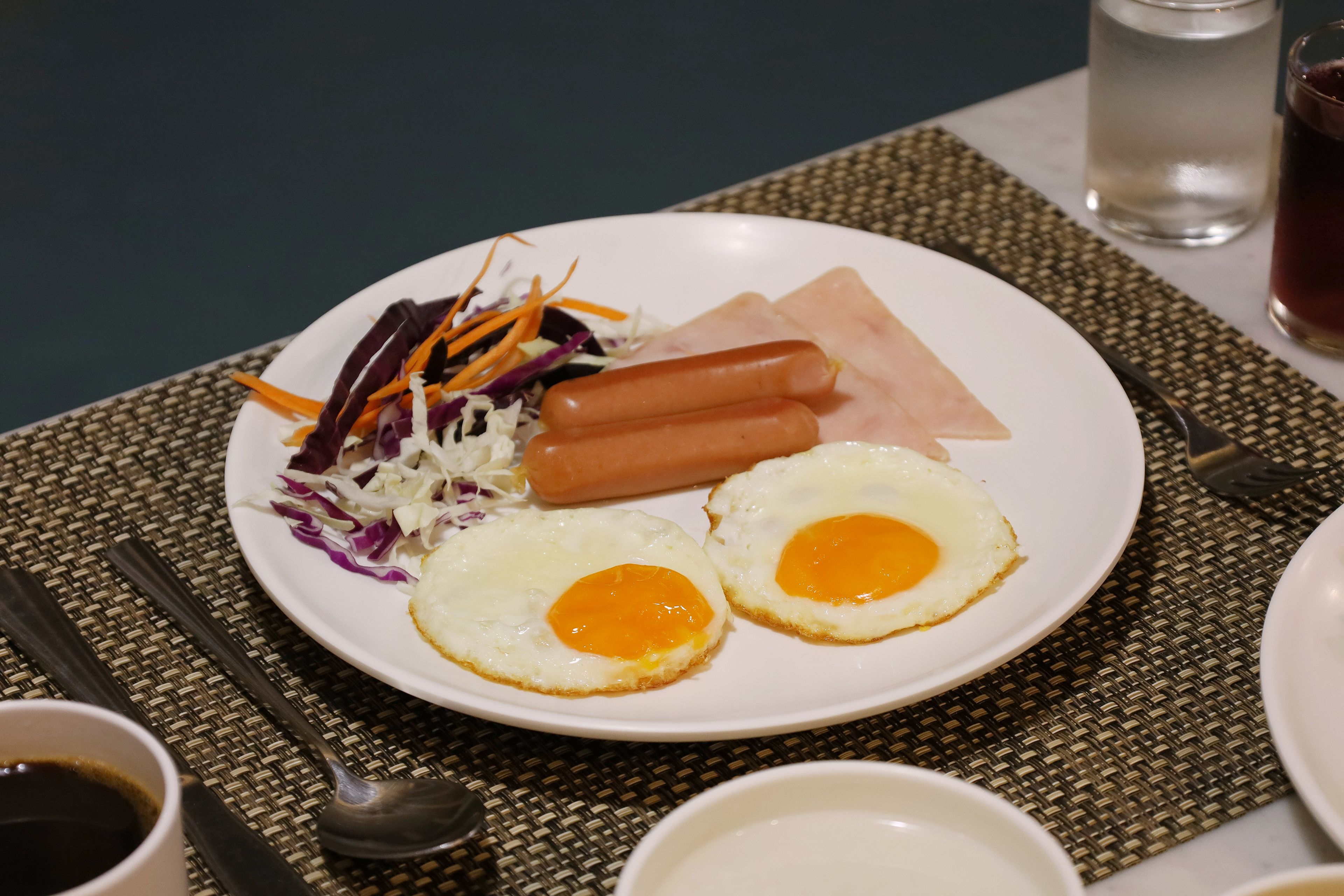 Un plato con dos huevos fritos, dos salchichas y una ensalada como guarnición sobre un mantel tejido.