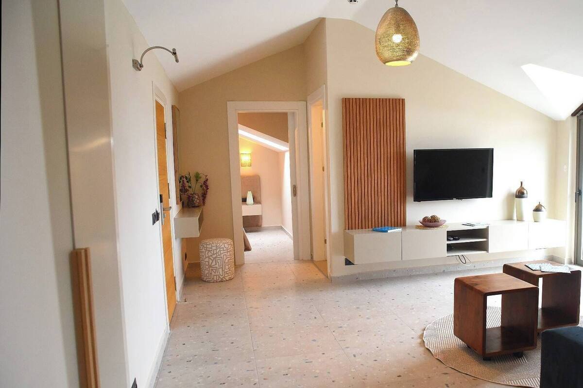Een ruime woonkamer met een aan de muur bevestigde televisie, een houten bankje en een gestreept voetenbankje.