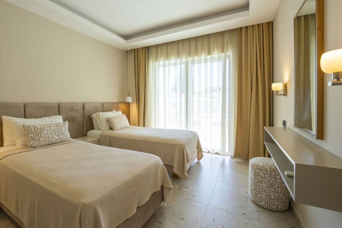 Een hotelkamer met twee bedden, nachtkastje, lamp, spiegel en een raam met gordijnen.