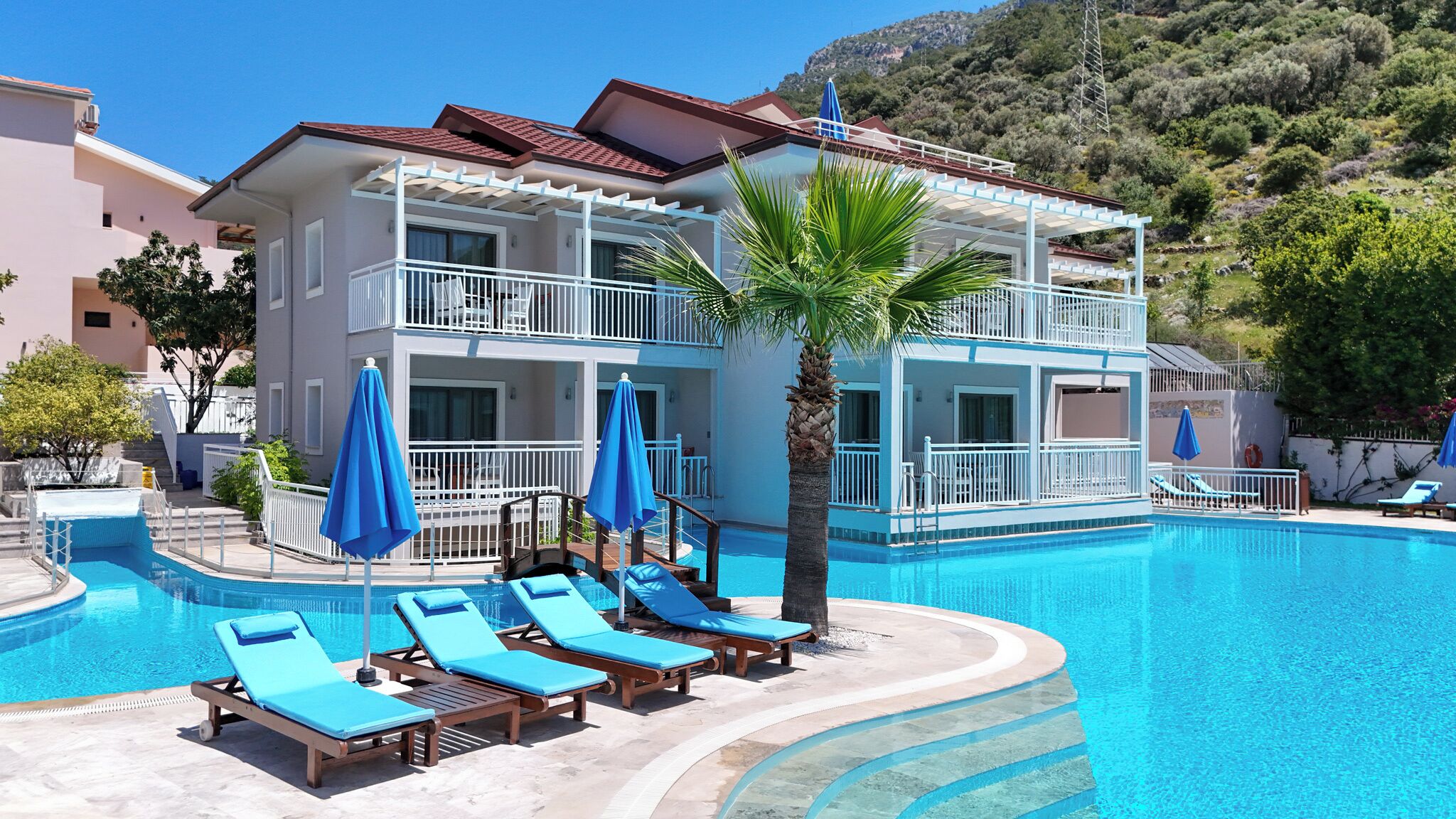 A two-story hotel with a swimming pool, sun loungers, and a palm tree.