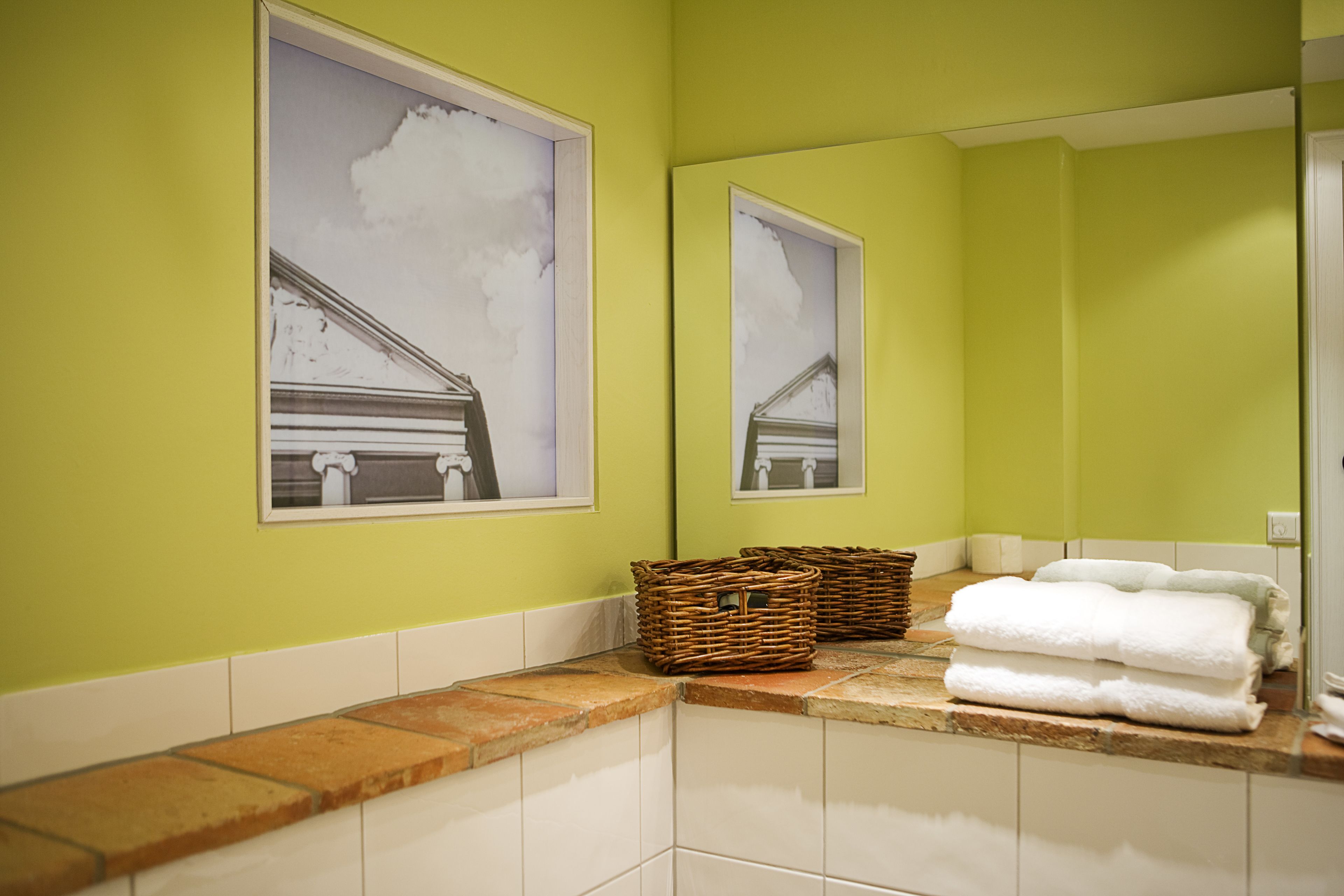 A bathroom with green walls, white tiles, and two framed pictures of a building. There are wicker baskets and folded towels on the counter.