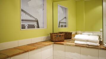 A bathroom with green walls, white tiles, and two framed pictures of a building. There are wicker baskets and folded towels on the counter.