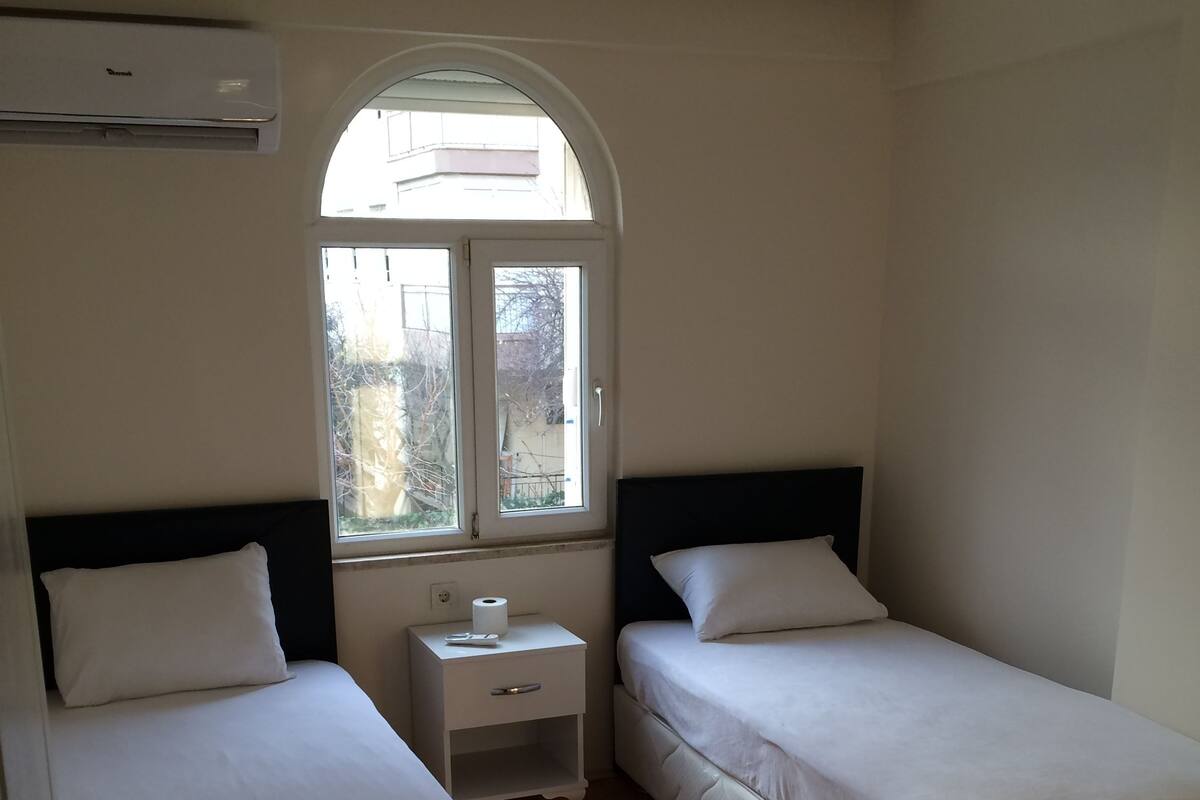 Une chambre avec deux lits simples, une petite table de chevet avec une lampe, une fenêtre donnant sur un beau paysage et un parquet en bois disposé en point de Hongrie.