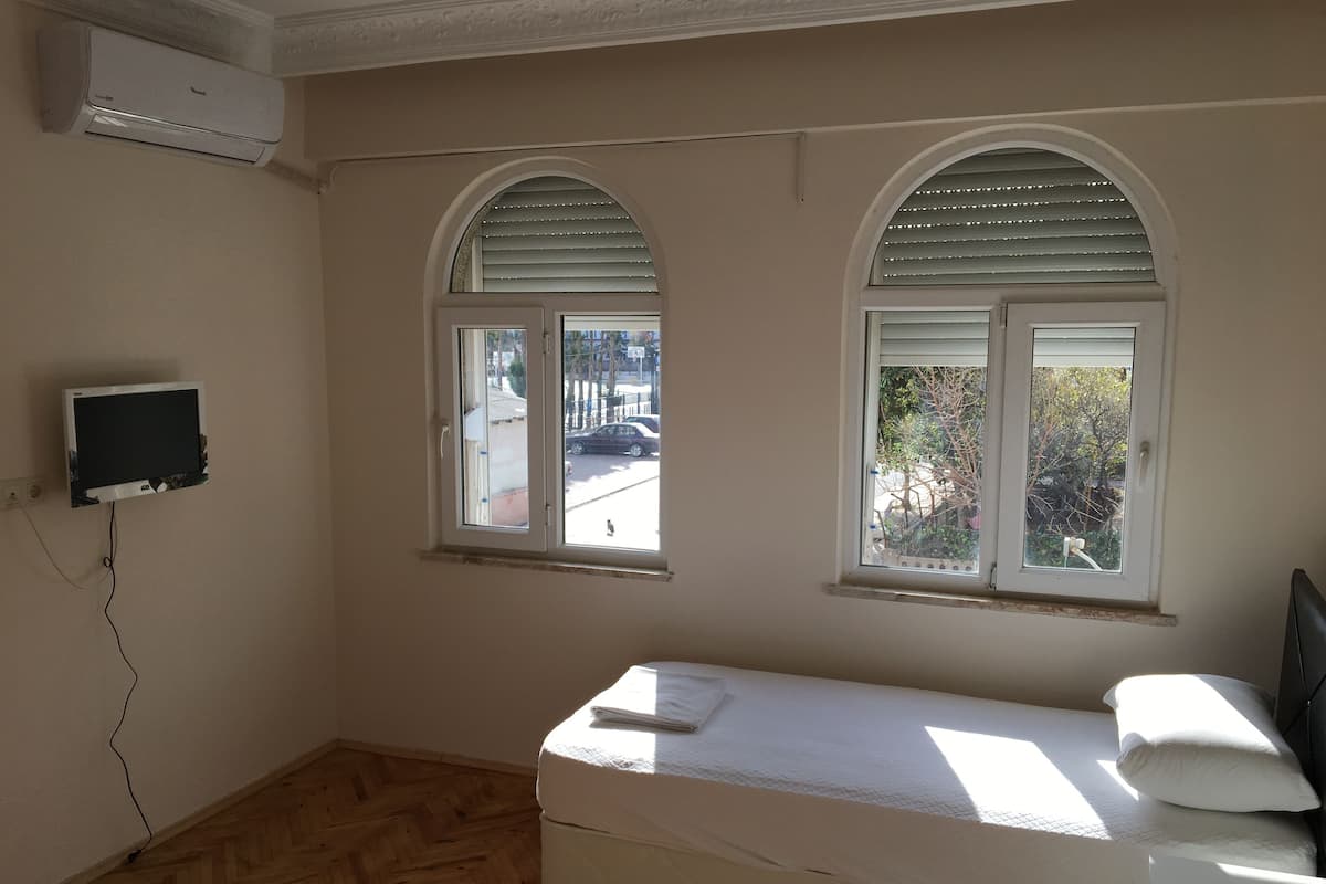 Une chambre avec un lit simple, deux fenêtres avec volets, un téléviseur fixé au mur et un climatiseur fixé au plafond.