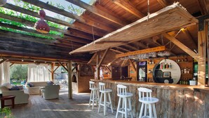 Un rústico área de bar con taburetes, una zona de estar con sofás, y vistas al exterior con vegetación.