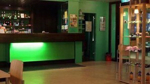 Zona de bar con mostrador iluminado en verde, área de estar con mesa y sillas, y vitrina con diversos artículos.