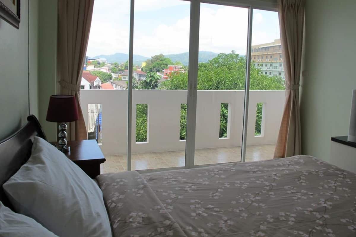 Ein Schlafzimmer mit Bett, Nachttisch, Lampe und Blick auf einen Balkon mit Geländer und Bepflanzung.