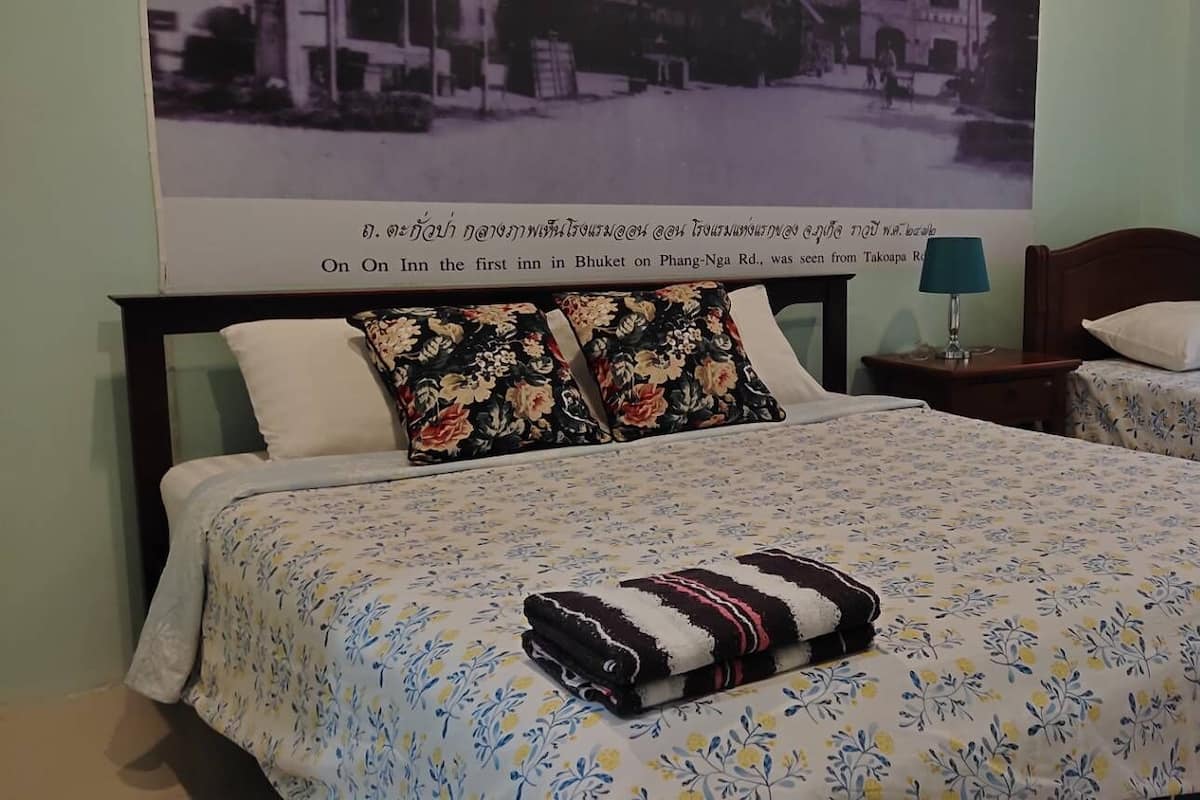 Ein Zimmer mit zwei Betten, einem größeren und einem kleineren, beide mit floralem Bettbezug. Zwischen den Betten steht ein Nachttisch mit einer Lampe, darüber an der Wand ein Poster.