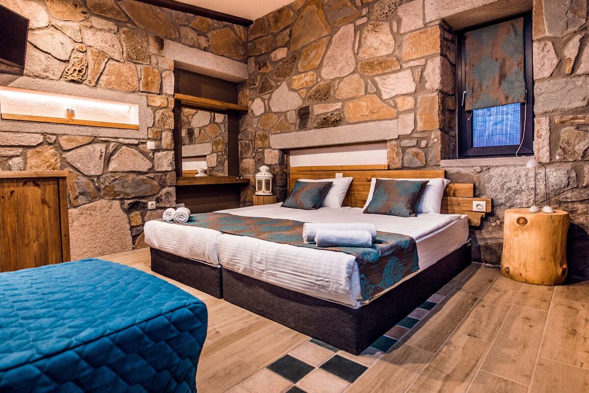 A bedroom with a stone wall, a large bed with multiple pillows, a wooden headboard, a nightstand with a lamp, and a small wooden stool.