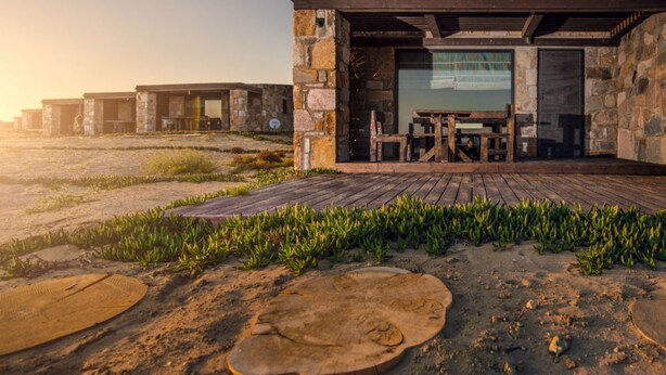 A rustic cabin with a wooden deck, stone walls, and a large window overlooking a grassy area. - Keros Blue (Lemnos)