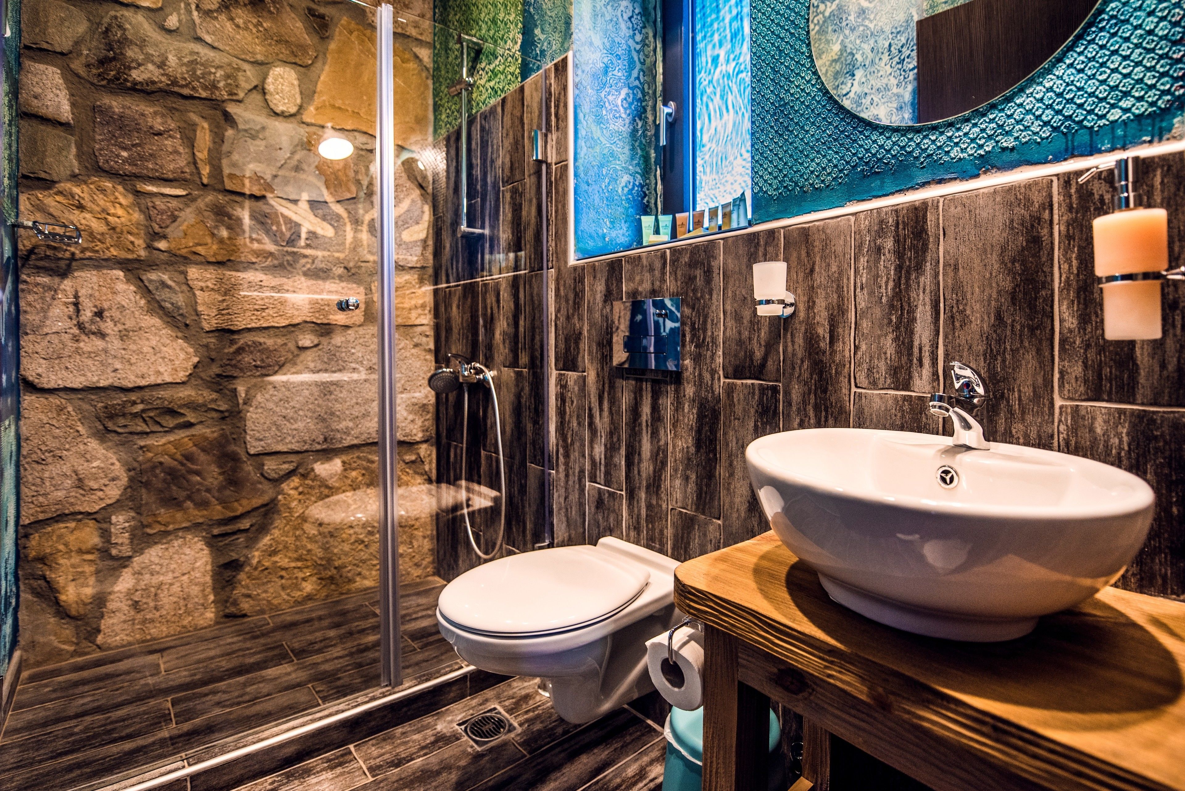 A bathroom with a stone wall, a wooden vanity, a white sink, and a glass shower enclosure.