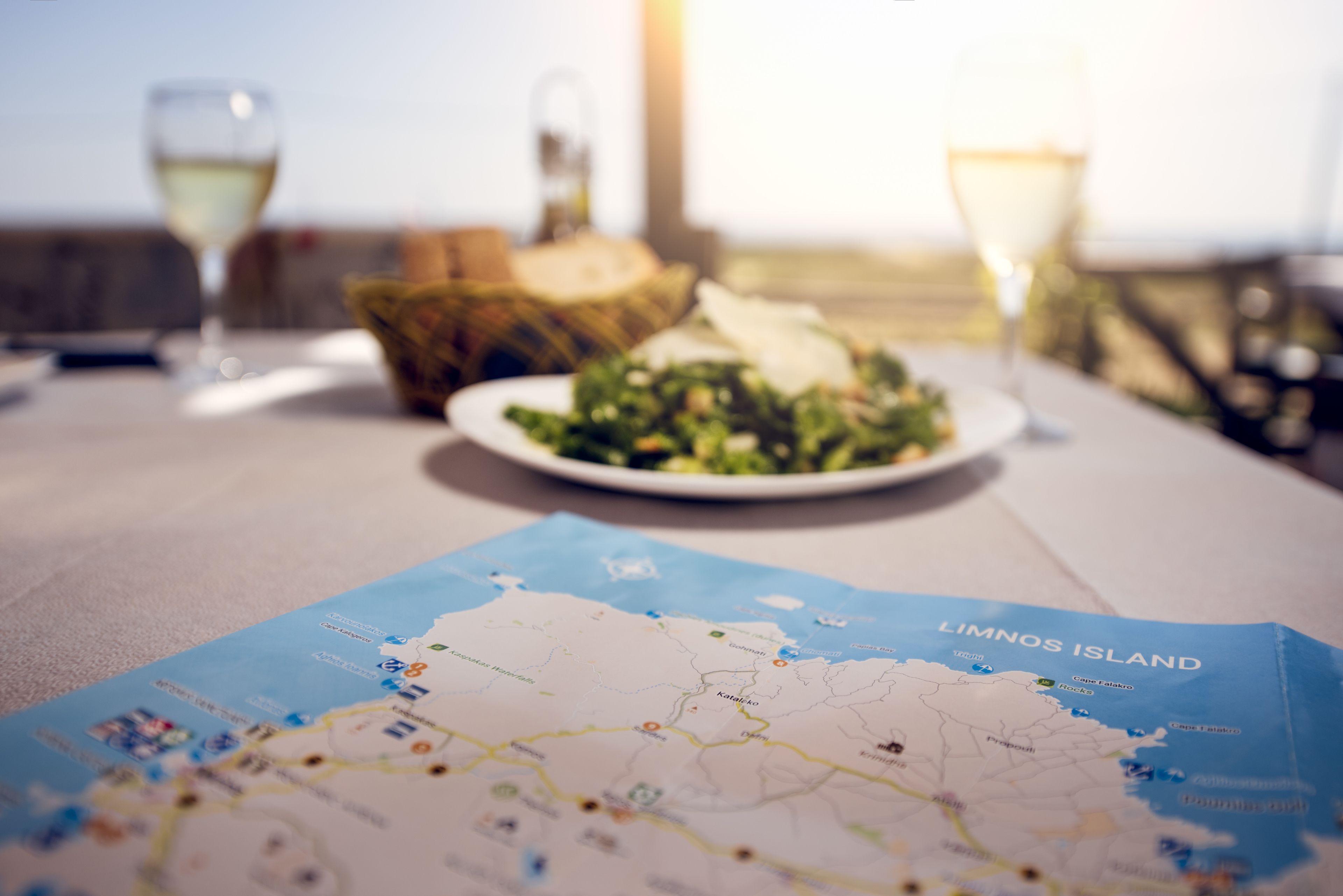 Una mesa de comedor con un mapa de la isla de Limnos, un plato de comida y dos copas de vino blanco.