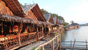 A row of thatched-roof overwater bungalows with wooden walkways over calm water. - Khaochan Resort (Pak Phayun)