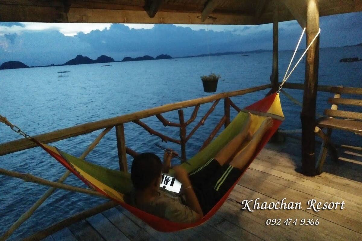 A hammock chair on a wooden deck overlooking a body of water with distant landforms.