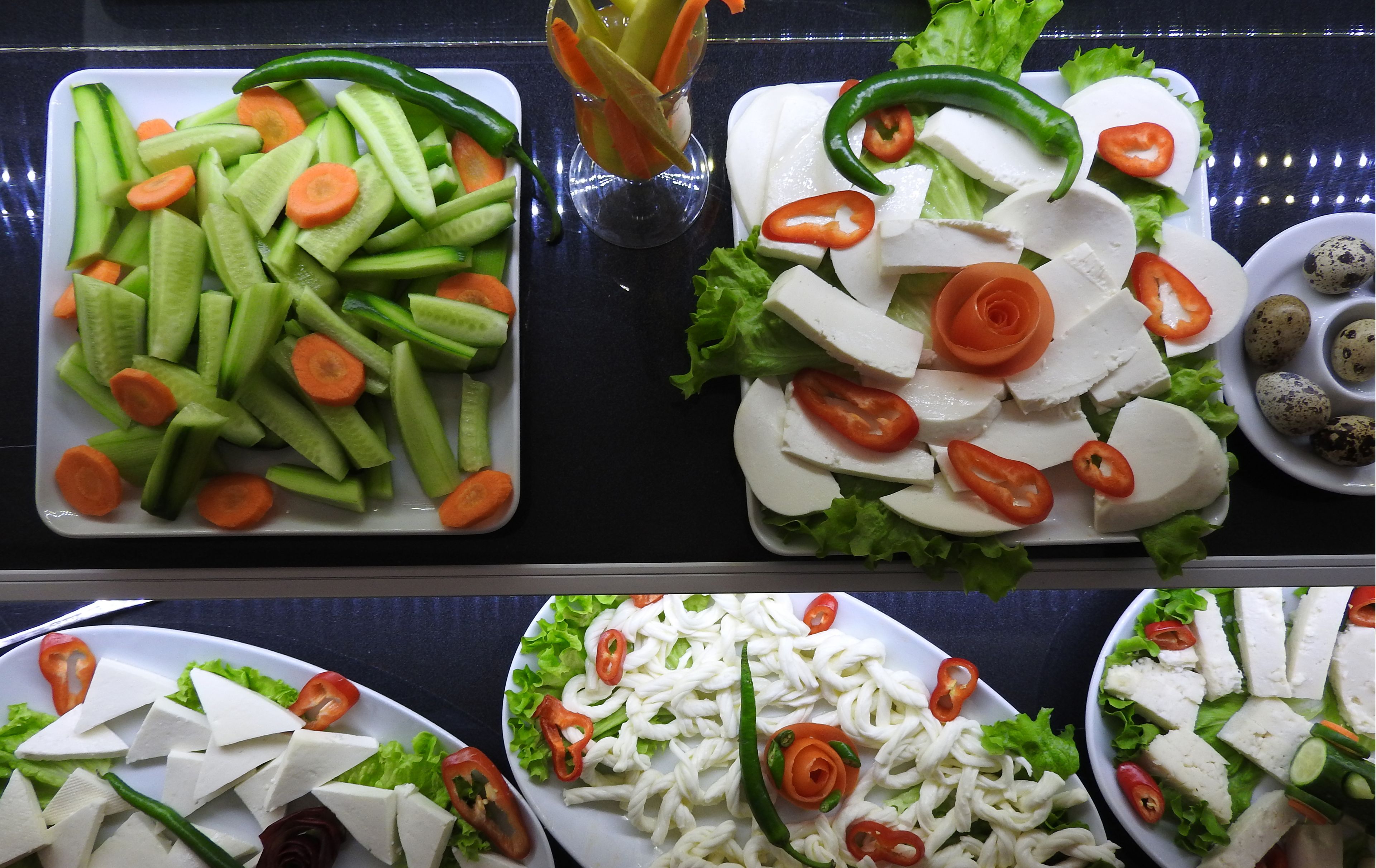 A buffet with various dishes including sliced cucumbers, carrots, tomatoes, lettuce, and cheese.
