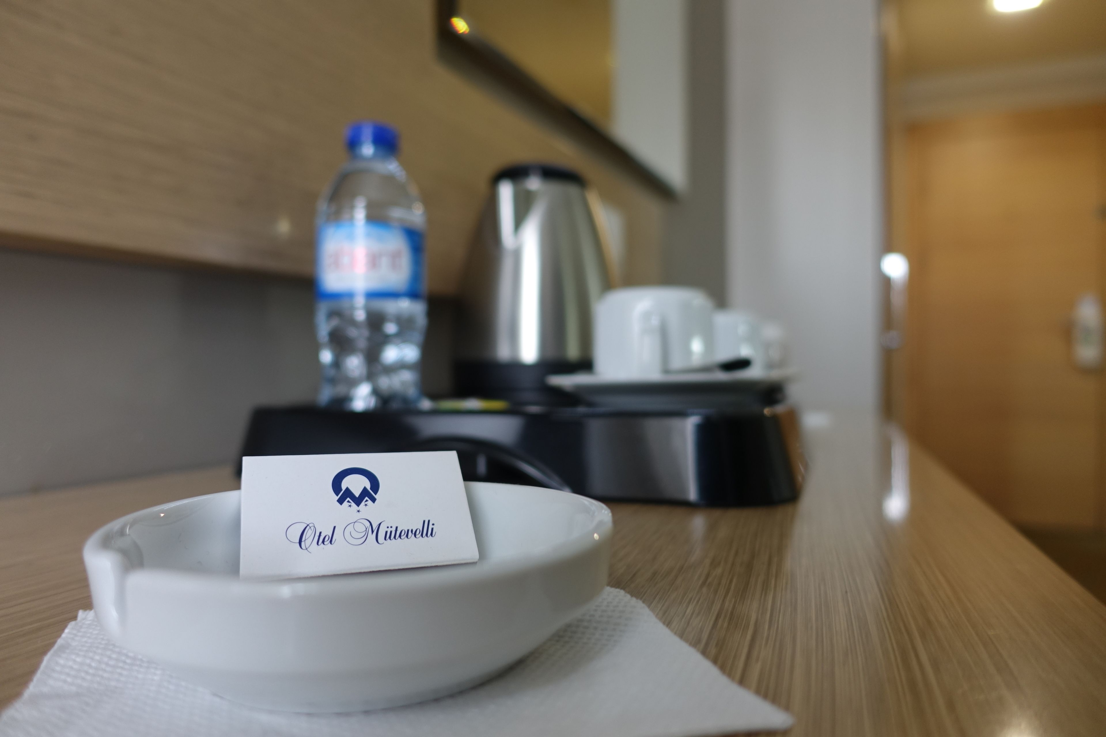 Una habitación de hotel con una mesa de madera, un bol blanco con una tarjeta de presentación, una botella de agua, una cafetera y una taza.