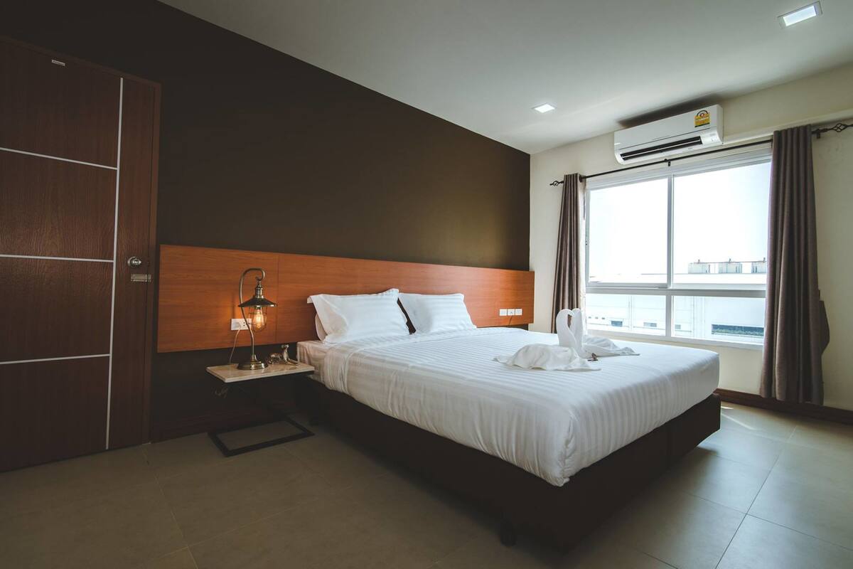Quarto de hotel com uma cama grande, uma mesa de cabeceira, uma luminária, uma janela com cortinas e um aparelho de ar condicionado.