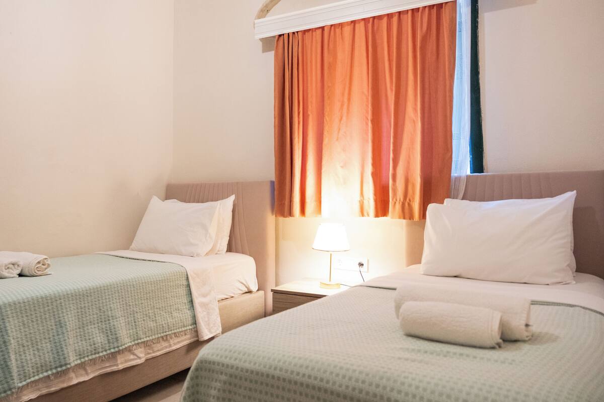 A hotel room with two single beds, each with a pillow and a rolled towel. There is a nightstand with a lamp between the beds.