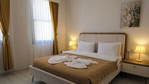 Cama bem arrumada com lençóis brancos e manta marrom xadrez, duas luminárias de cabeceira e uma pintura floral na parede.