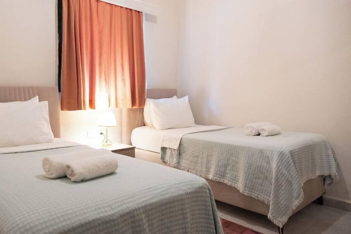 A hotel room with two beds, each with white bedding and pillows, a nightstand with a lamp, and a window with curtains.