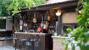 Uma área de bar ao ar livre com mobiliário de madeira rústica, variedade de garrafas e copos em prateleiras, e luzes decorativas penduradas.
