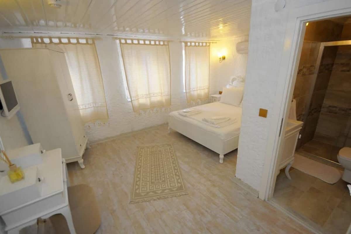 A room with a bed, a small desk, a chair, a window with blinds, and a bathroom with a shower and toilet.