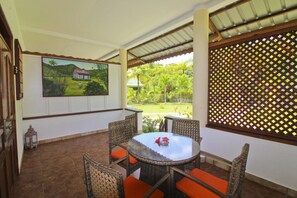 A covered patio with wicker chairs and orange cushions, a round table, and a painting of a rural scene.