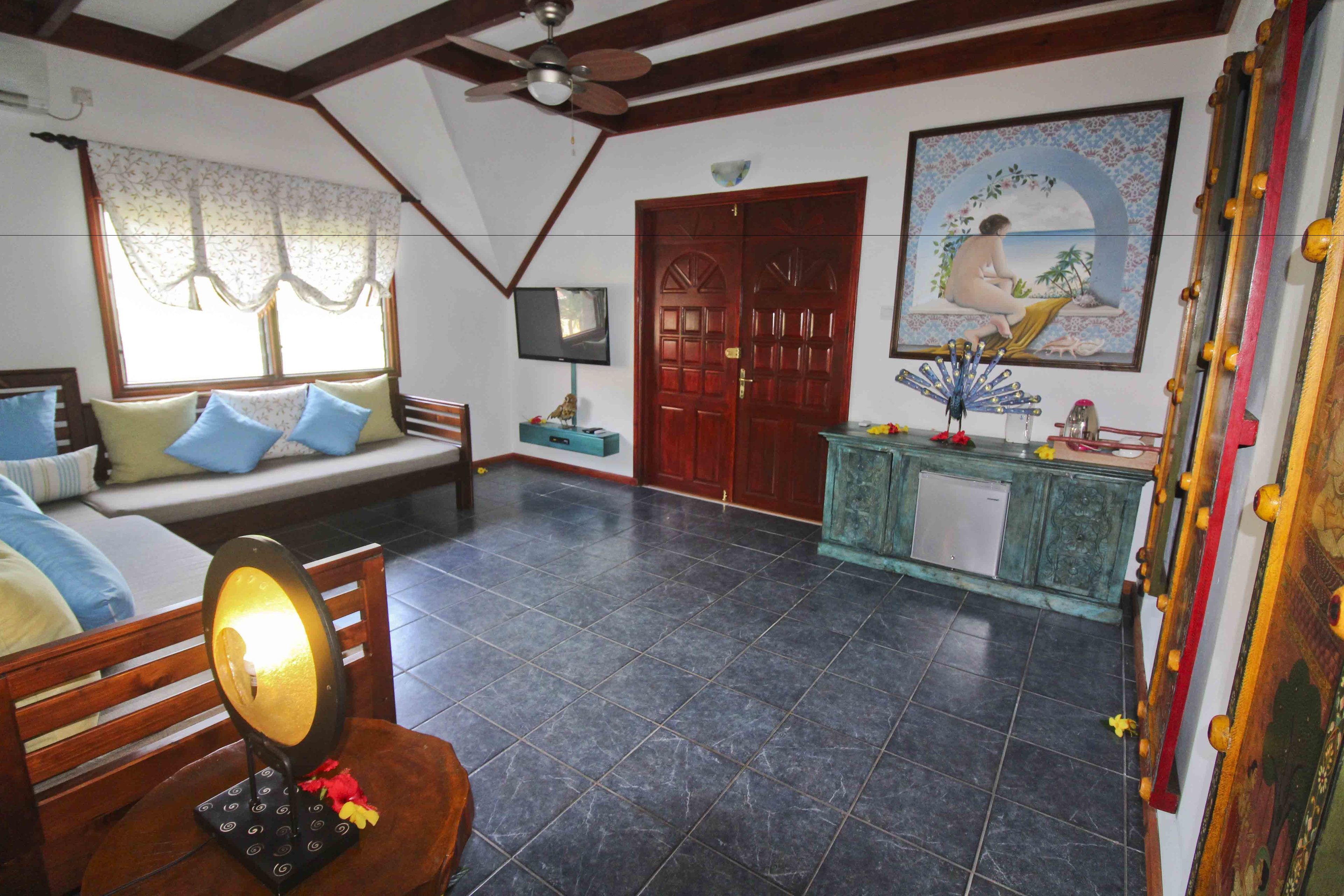 A room with a wooden ceiling, a ceiling fan, a television on the wall, a wooden door, a painting, and a tiled floor.