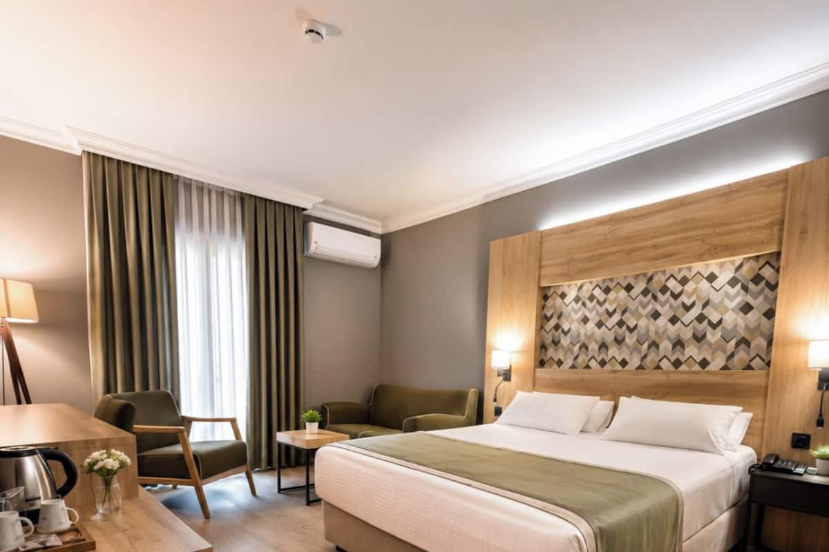 A hotel room with a bed, a desk with a lamp, a chair, and a wall-mounted air conditioner.