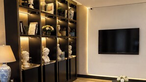 A modern living room with a large black bookshelf, a flat-screen TV mounted on the wall, a round armchair, and a coffee table with a book and vase. - Laodikya Otel (Denizli)