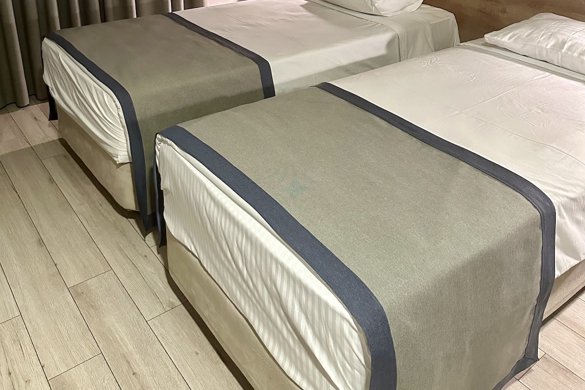 A hotel room with two beds, a wooden headboard, a nightstand with a lamp, and a window with blinds.