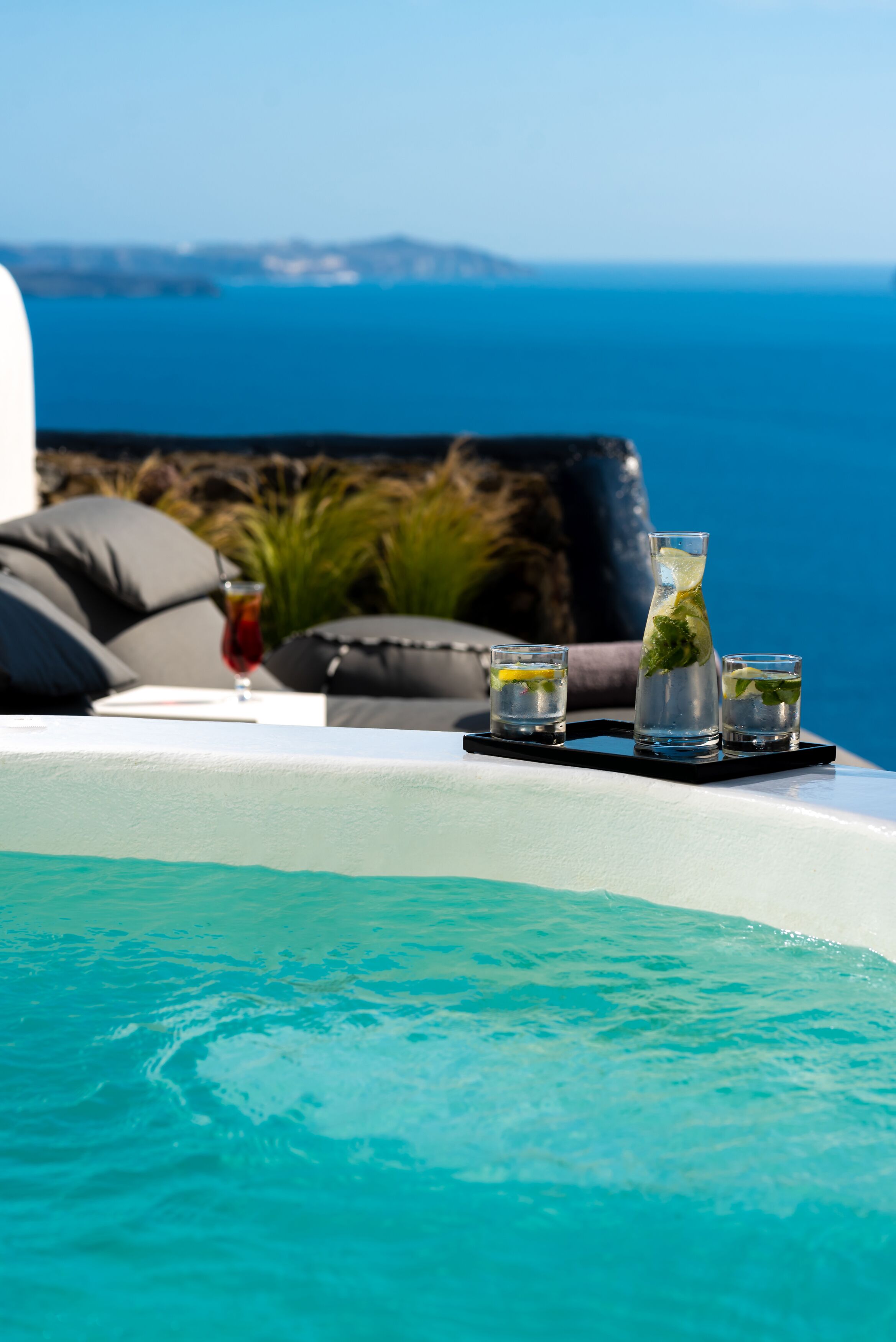 Une scène au bord de la piscine, avec un plateau contenant trois boissons, une table blanche et une vue sur l’océan et la côte lointaine.