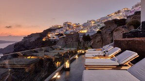 Una terraza en la azotea con sillones de descanso y barandilla de cristal, con vistas a un pueblo costero al atardecer.