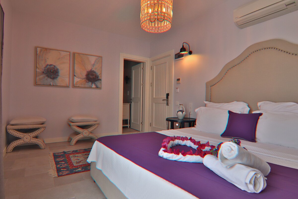 A bedroom with a bed, two bedside tables, a nightstand, a chandelier, and a door leading to another room.