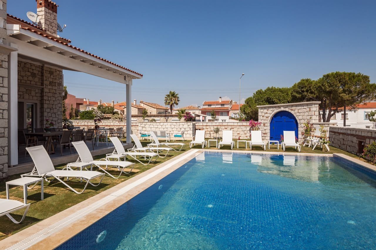 A swimming pool with white lounge chairs, a stone building with a blue door, and a clear blue sky.