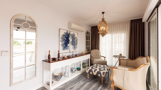 A living room with a white console table, a beige armchair, a patterned ottoman, a chandelier, and a large window with curtains. - Alacaat Otel (Cesme)
