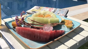 A poolside setting with a table holding a fruit platter, lounge chairs, and a stone building in the background. - Alacaat Otel (Cesme)