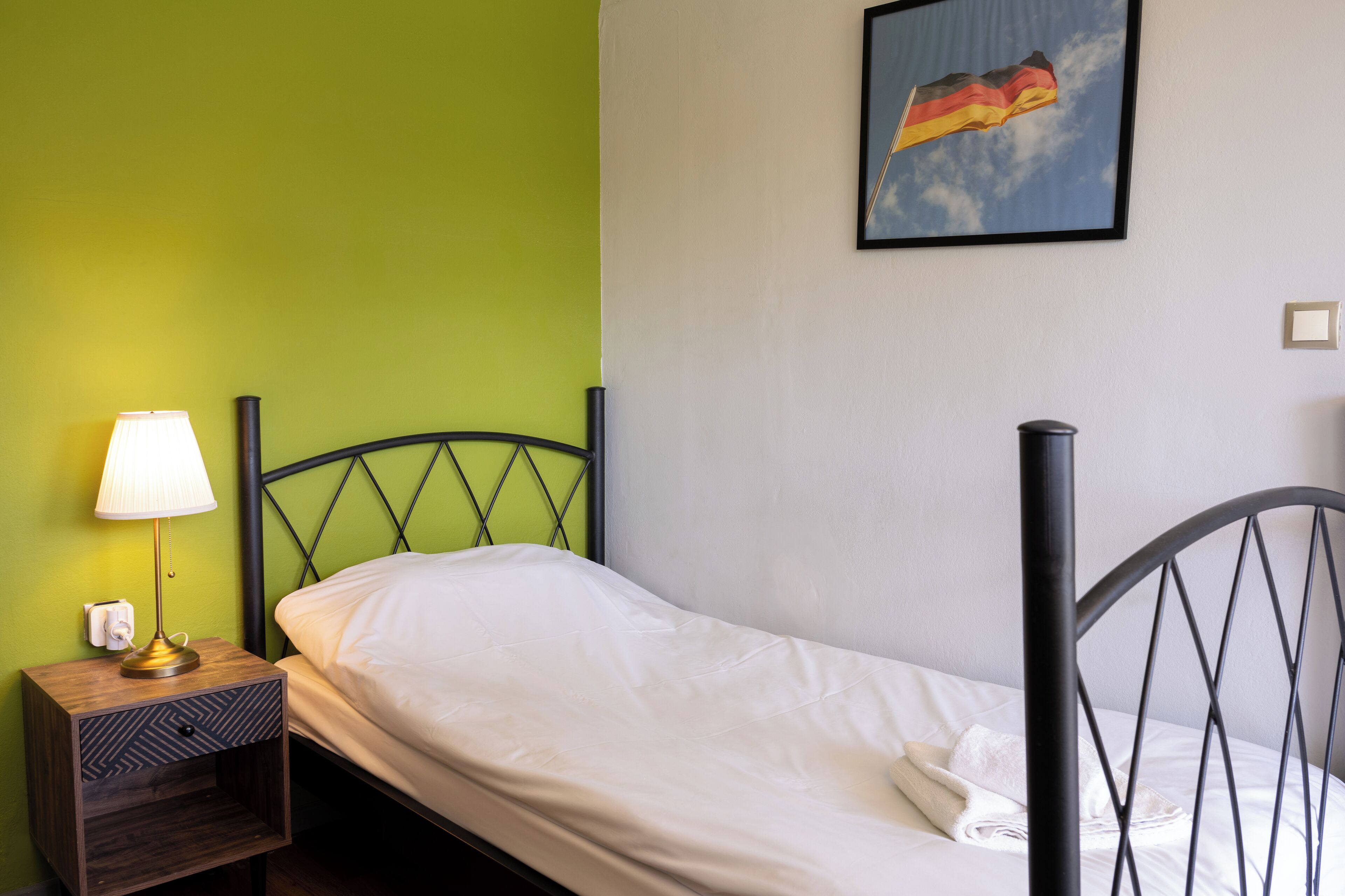 Una cama individual con cabecera, una mesita de noche con lámpara y un cuadro colgado en la pared con una bandera.