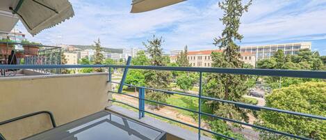 Ein Balkon mit Glastisch und Stühlen, einem blauen Geländer und Blick auf Gebäude und Bäume.