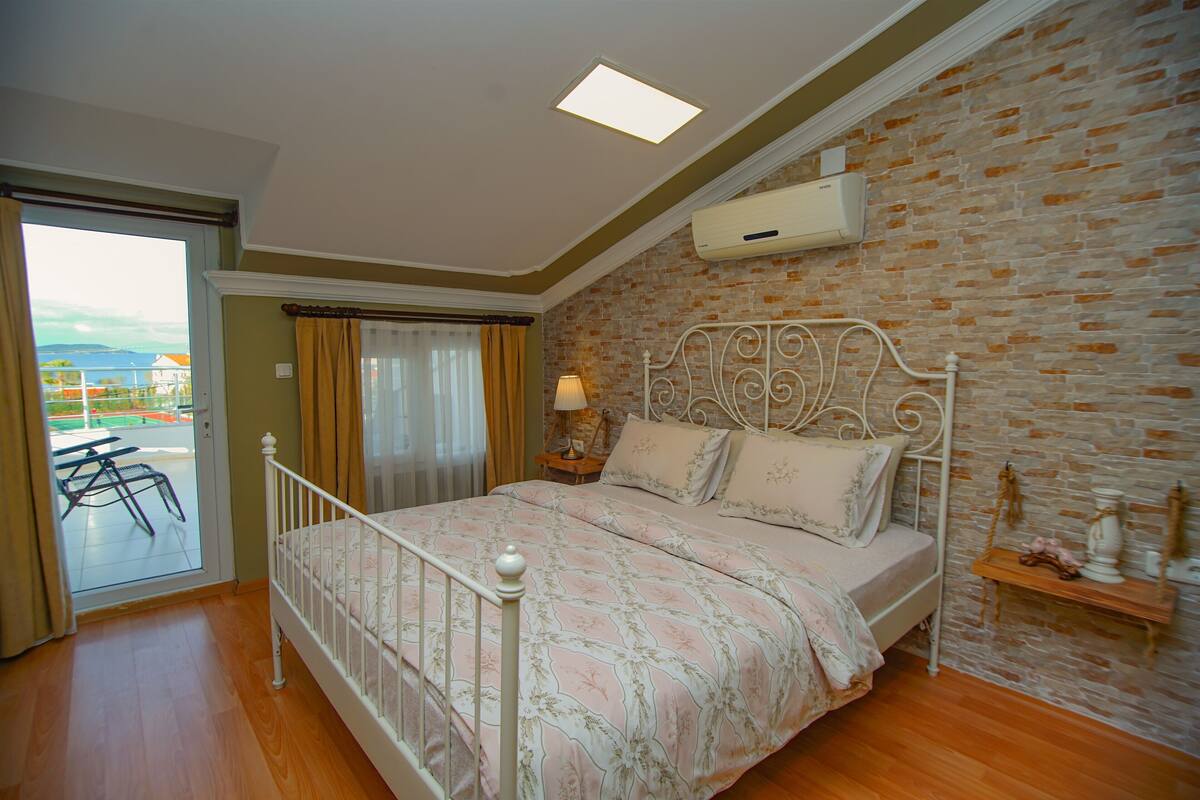 A bedroom with a brick wall, a bed with a metal frame, a window with curtains, and a view of the outdoors.