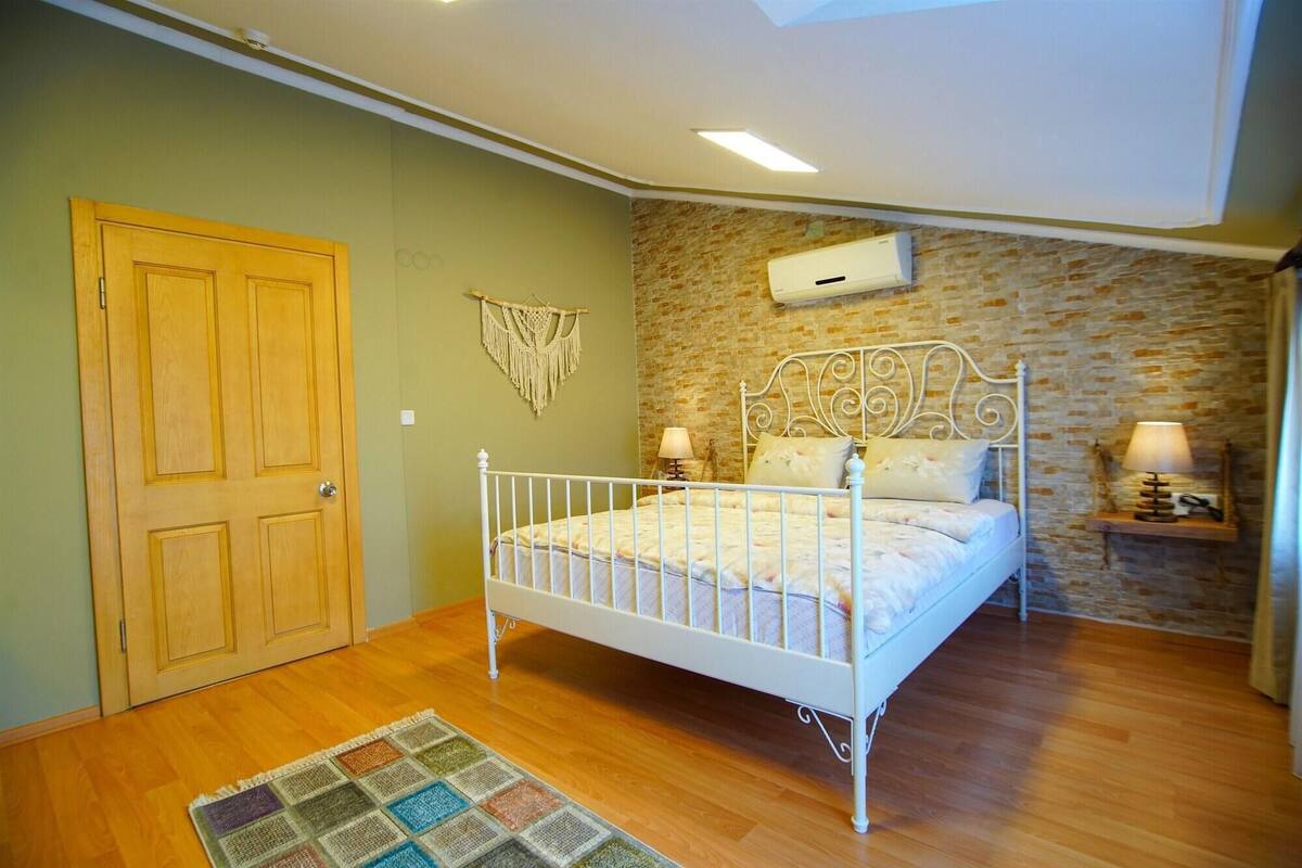 A bedroom with a wooden door, a white metal bed, a stone wall, and a small bedside table with a lamp.