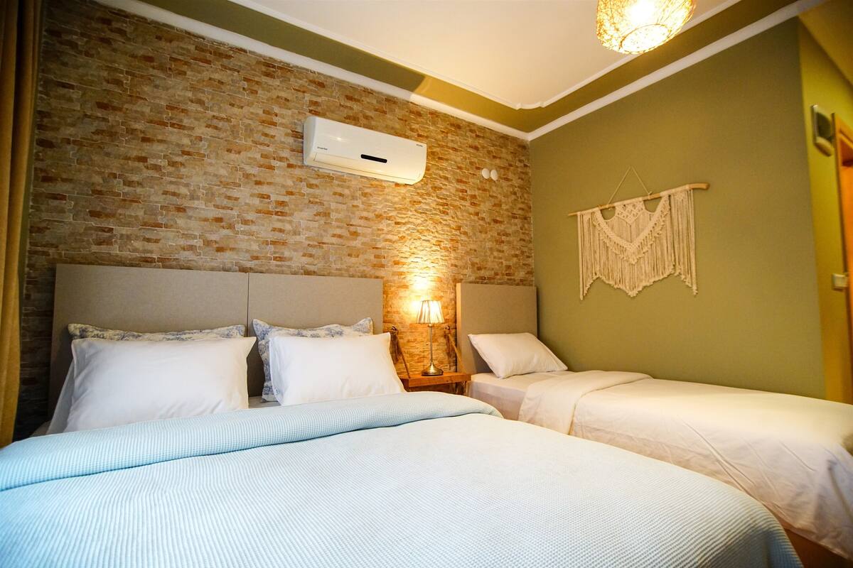 A hotel room with a brick wall, a bed with white bedding, a bedside lamp, and a wall-mounted air conditioner.