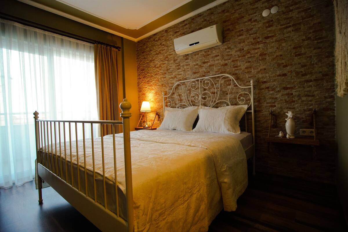 A bedroom with a brick wall, a bed with white bedding, a nightstand with a lamp, and a window with curtains.