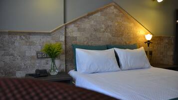 Una habitación de hotel con pared de ladrillo decorativa, una cama con ropa blanca, una mesita de noche con lámpara, y un jarrón con flores.