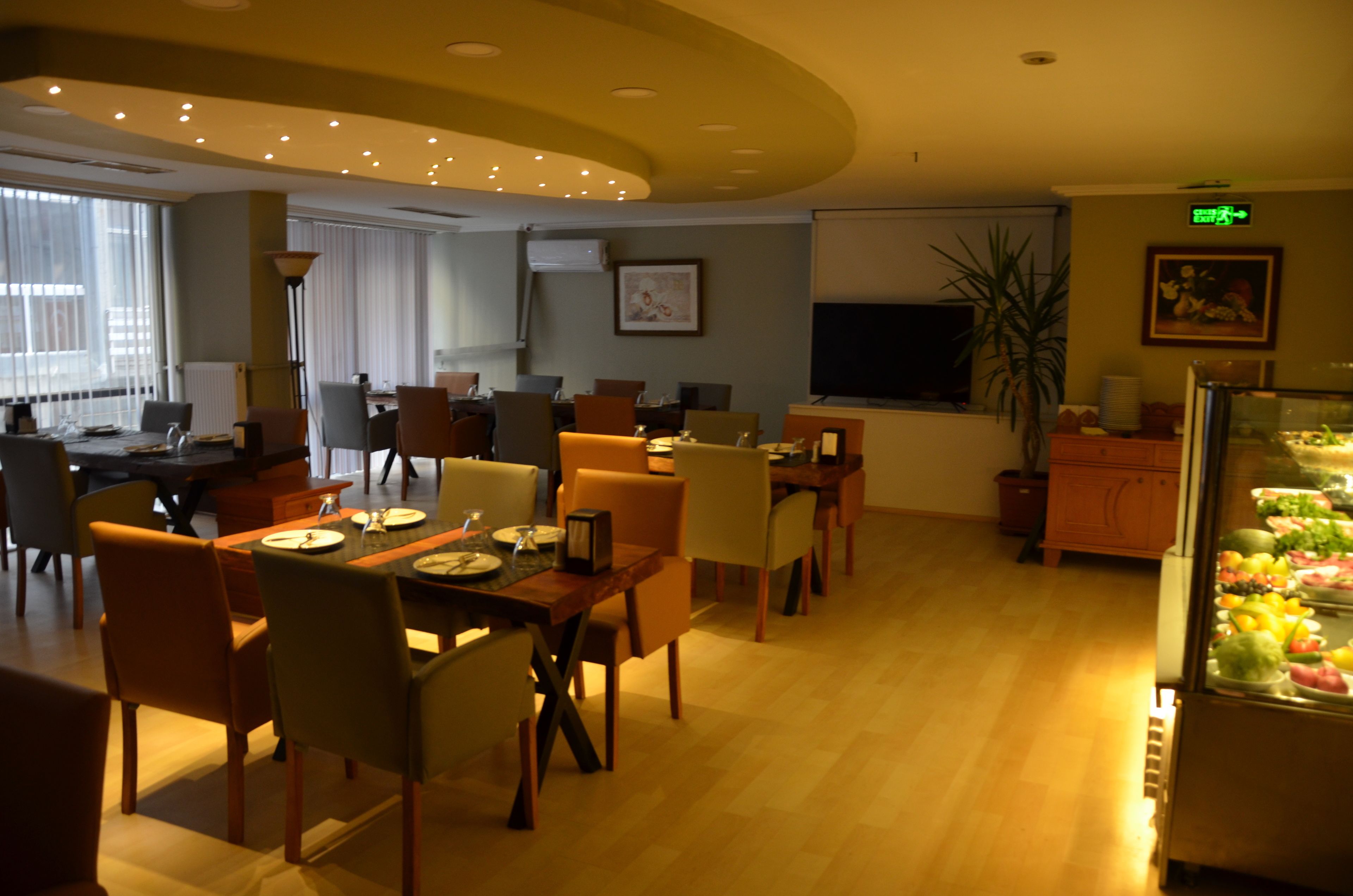 Une salle à manger d’hôtel avec plusieurs tables et chaises, un présentoir de fruits et un écran de télévision fixé au mur.