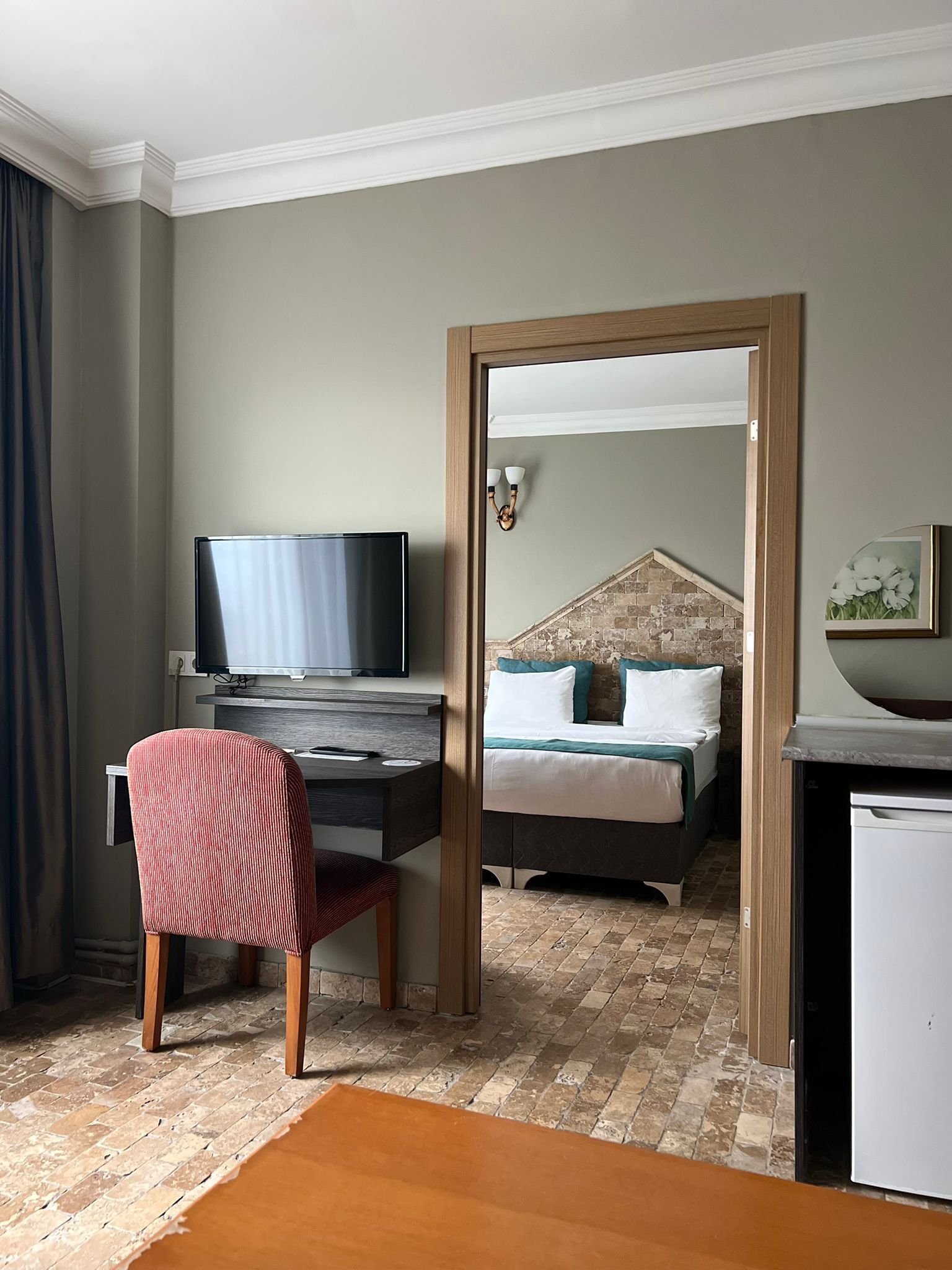 A hotel room with a bed, a desk with a TV, a chair, and a small refrigerator.