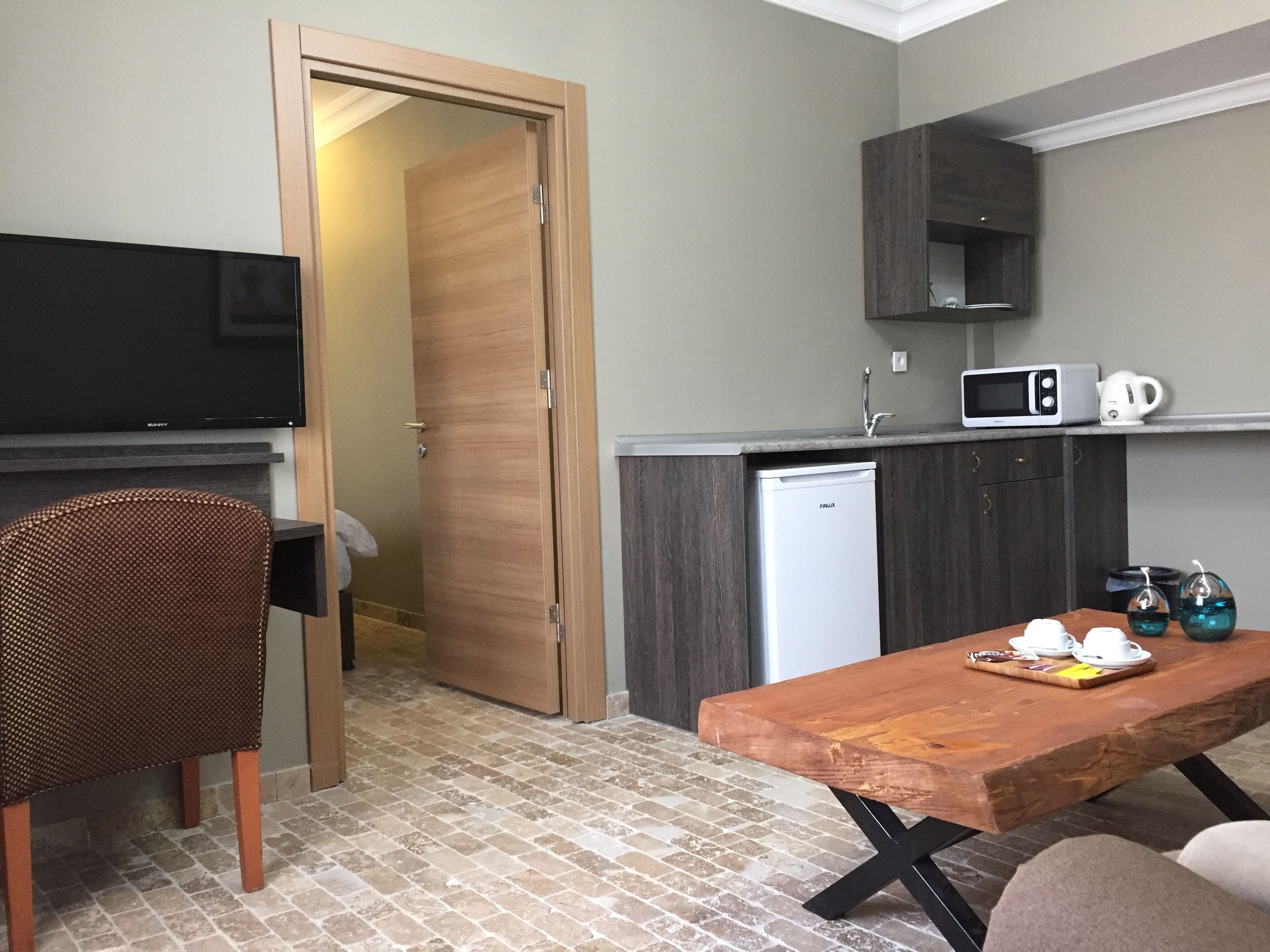 Eine kompakte Küche mit Mikrowelle, Wasserkocher und kleinem Kühlschrank. Ein Holztisch mit einer Tablett voller Tassen und einem Stuhl.